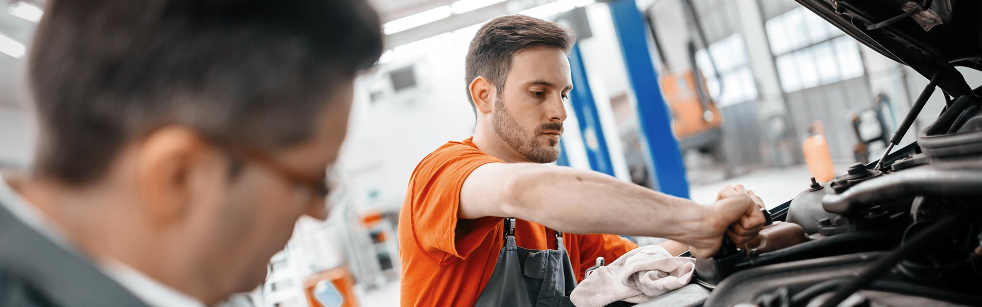 A mechanic working on a car engine while another person observes in a workshop.